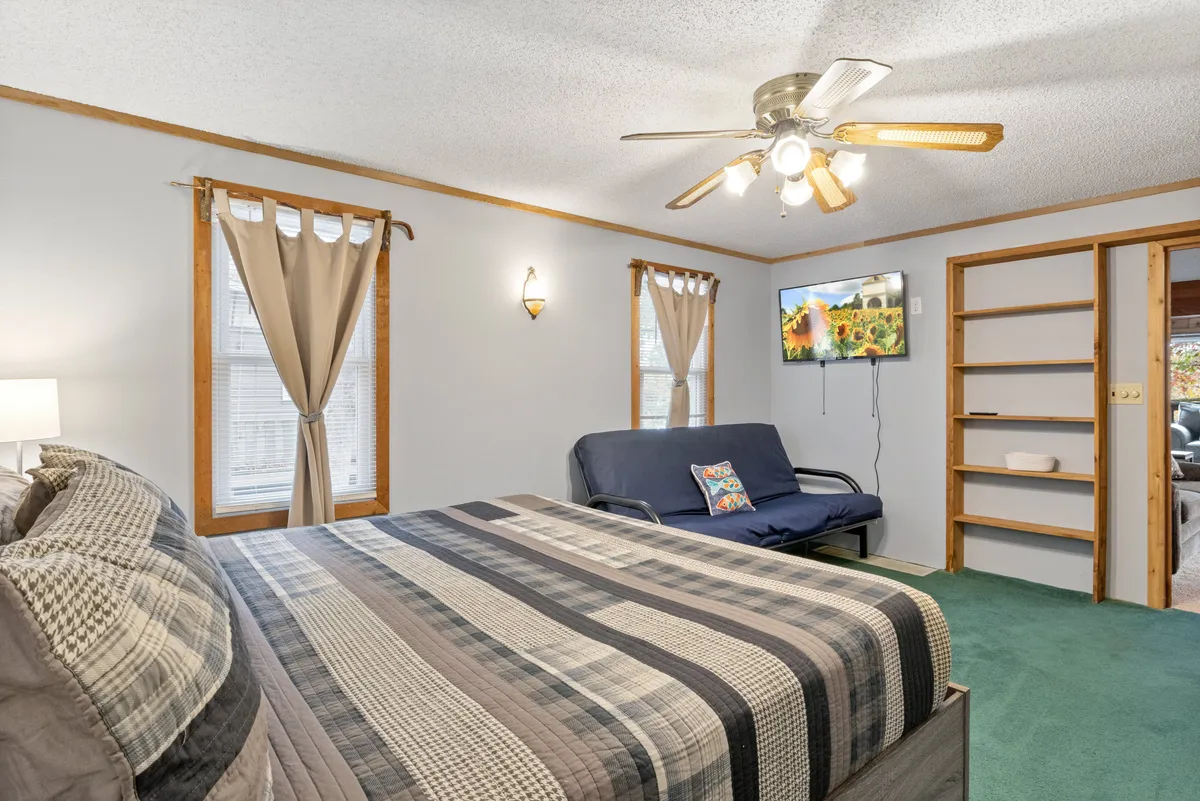 A bright bedroom with a bed, sofa, TV, and windows that let in sunlight. The arrangement provides both a snug sleeping area and a spot for relaxing or watching movies, catering to different guest needs.