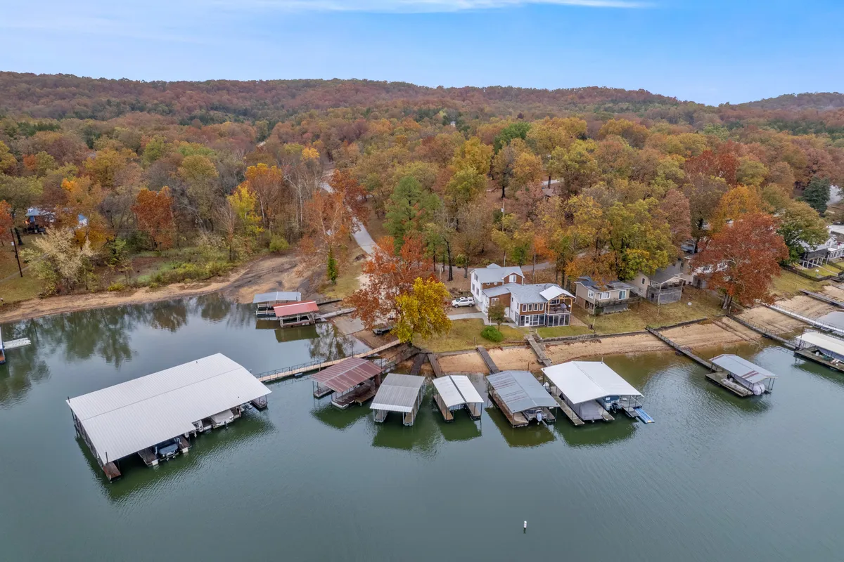 A serene lakeside scene featuring the lake, boathouses, autumn foliage, and houses dotting the shoreline. The water is calm, with a few boats in the distance, and golden leaves frame the edges of the view.
