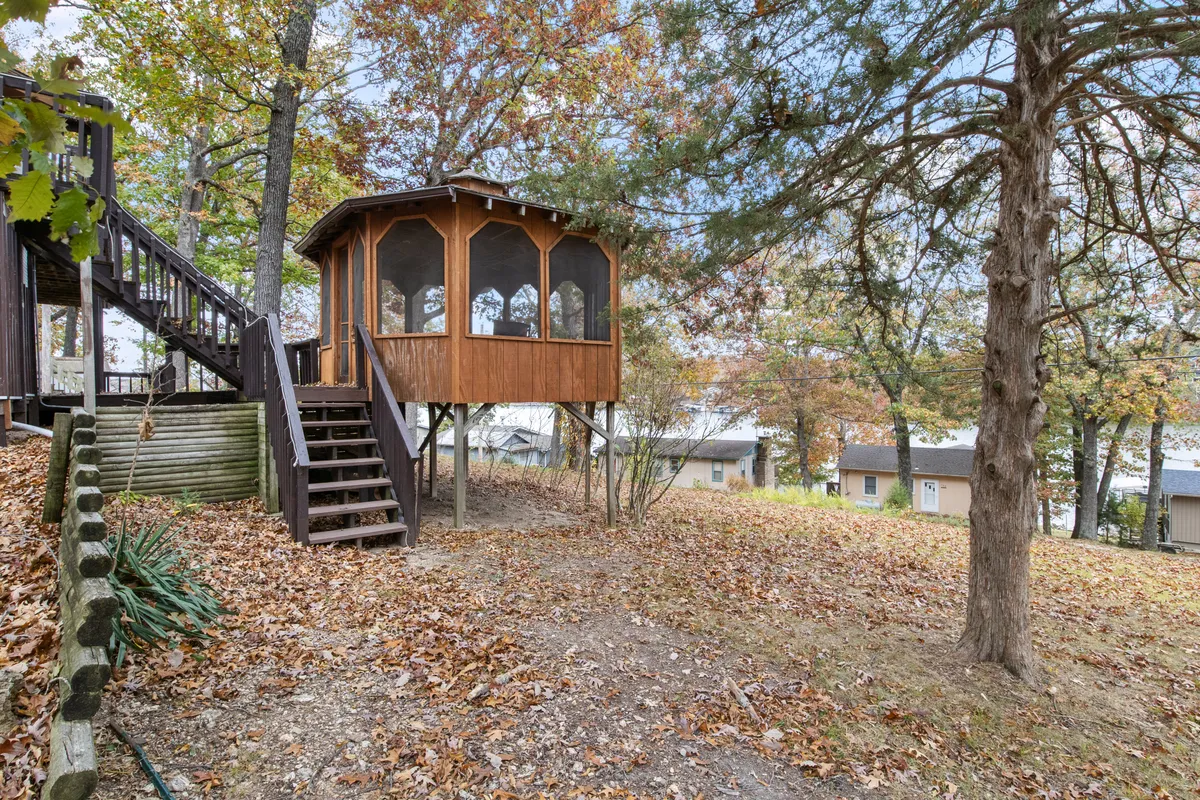An elevated gazebo nestled among leafy trees with a ground scattered in autumn leaves, inviting guests to relax, enjoy nature views, or gather socially in a peaceful outdoor setting.