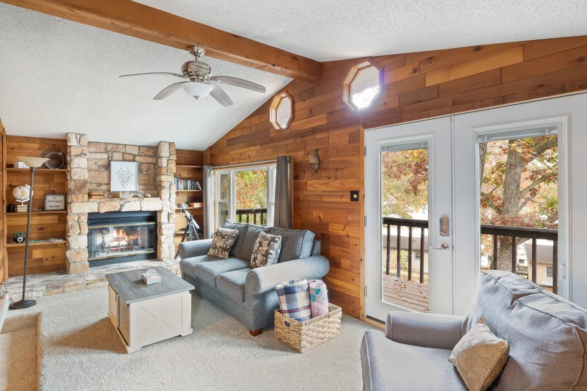 A cozy living area with a stone fireplace, a comfortable sofa, large windows that let in natural light, and warm wooden walls. The windows offer glimpses of the outdoors, enhancing the inviting atmosphere and highlighting the connection to the lake beyond.
