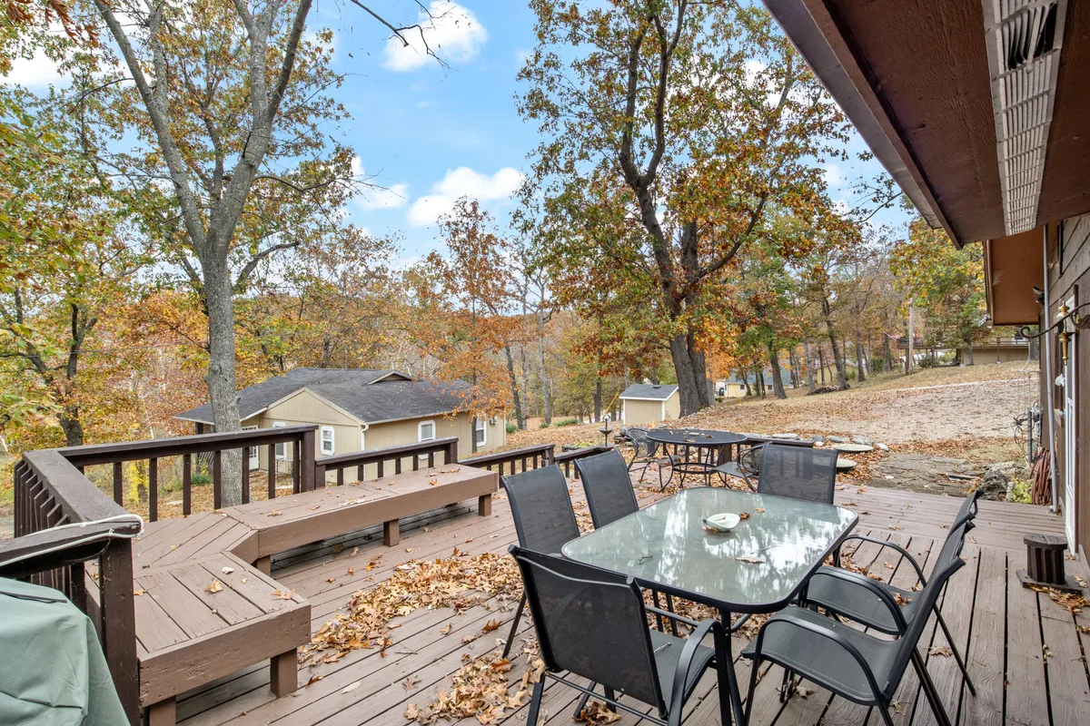 An inviting wooden deck is set with a glass dining table surrounded by lush trees and vibrant fall foliage, creating an intimate setting for outdoor meals. The space radiates tranquility and offers a beautiful vantage point for enjoying changing seasons and sunset views.