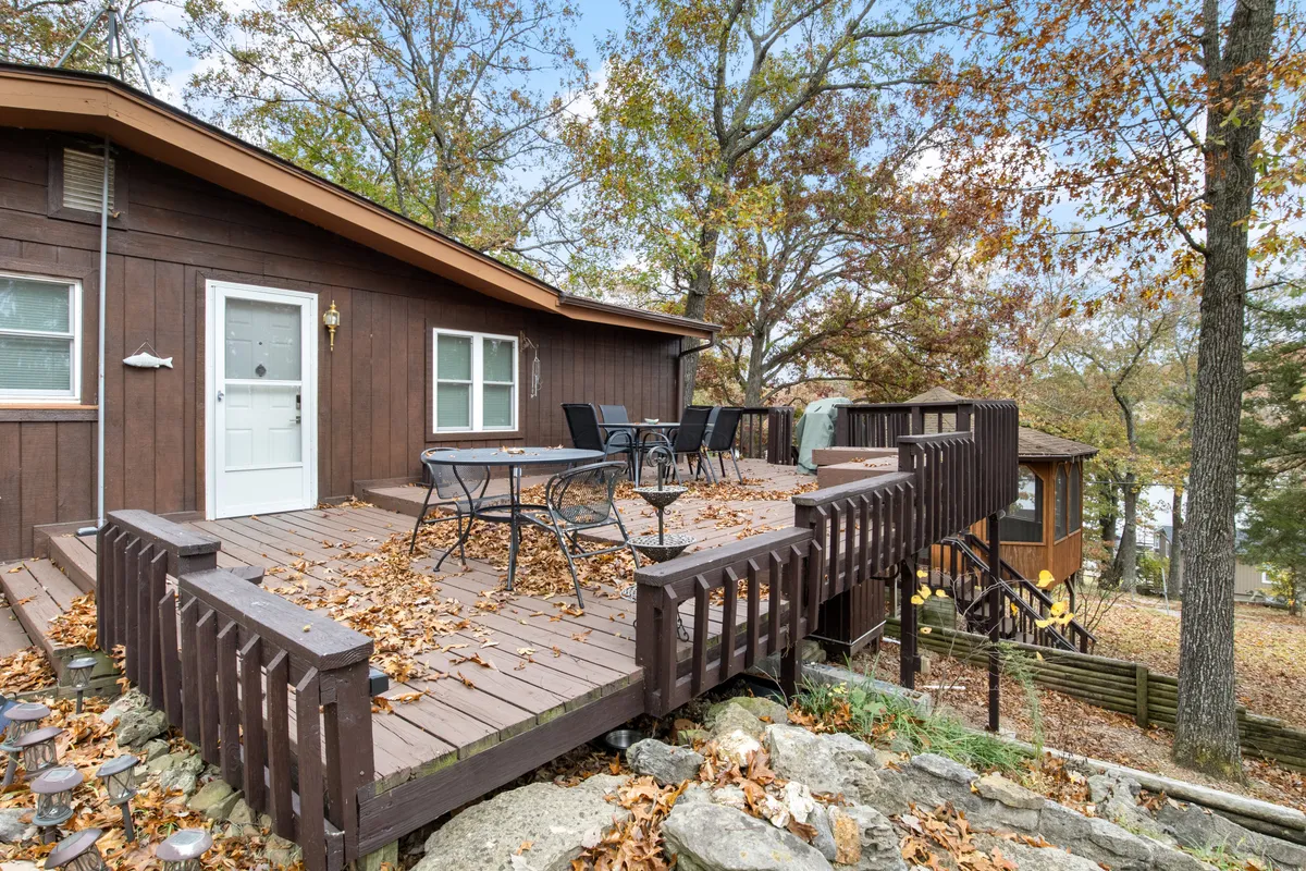A spacious wooden deck surrounded by mature trees, featuring a large outdoor dining table and comfortable chairs. Steps lead down to a lower area, and the space is bathed in natural light, giving a welcoming and open feel ideal for gatherings or quiet relaxation.