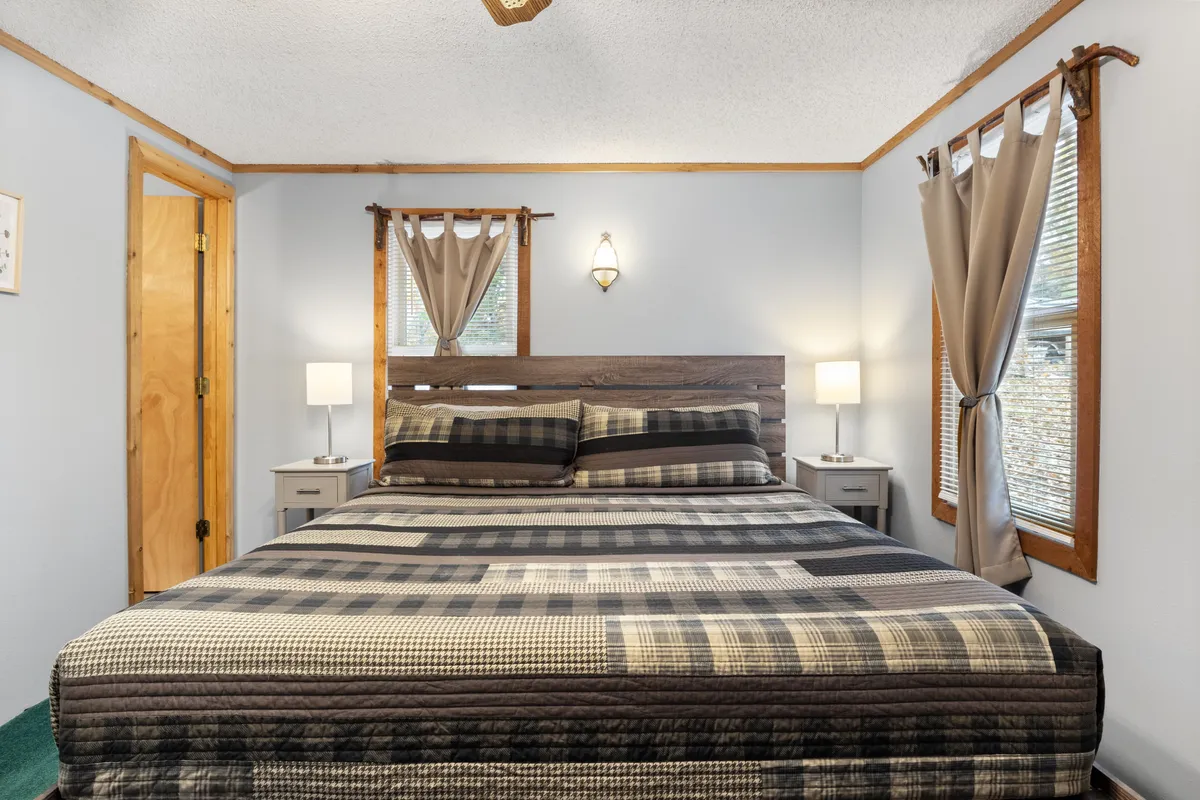 A cozy bedroom featuring a bed with patterned bedding, a sturdy wooden bed frame, nightstands with lamps on either side, and windows adorned with curtains that allow natural light to filter in. The setting feels peaceful and inviting, perfect for unwinding at the end of the day.