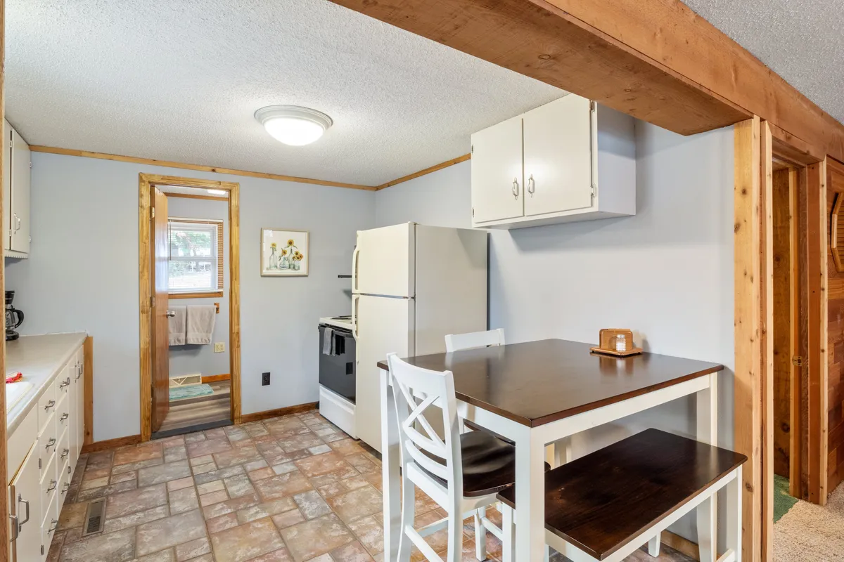 A homey dining area with a central table, surrounded by kitchen cabinets and a refrigerator, with a doorway leading into another space. The setting suggests shared meals, cooking together, and seamless flow between spaces.
